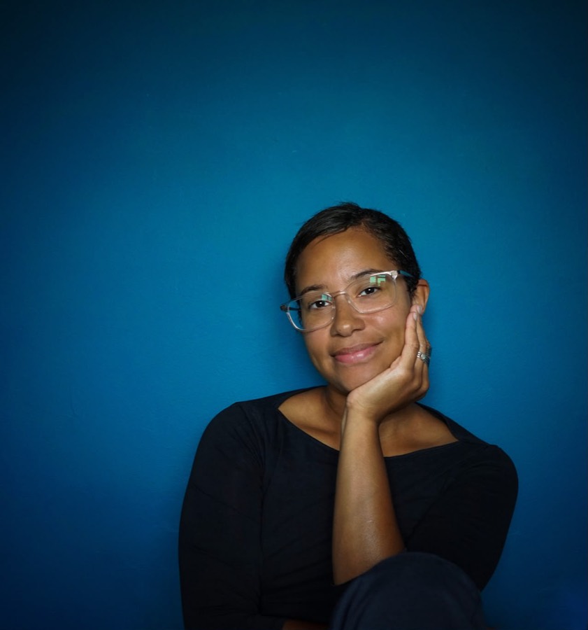 Jenyfa Martin, smiling with her chin resting on her hand against a deep blue wall.