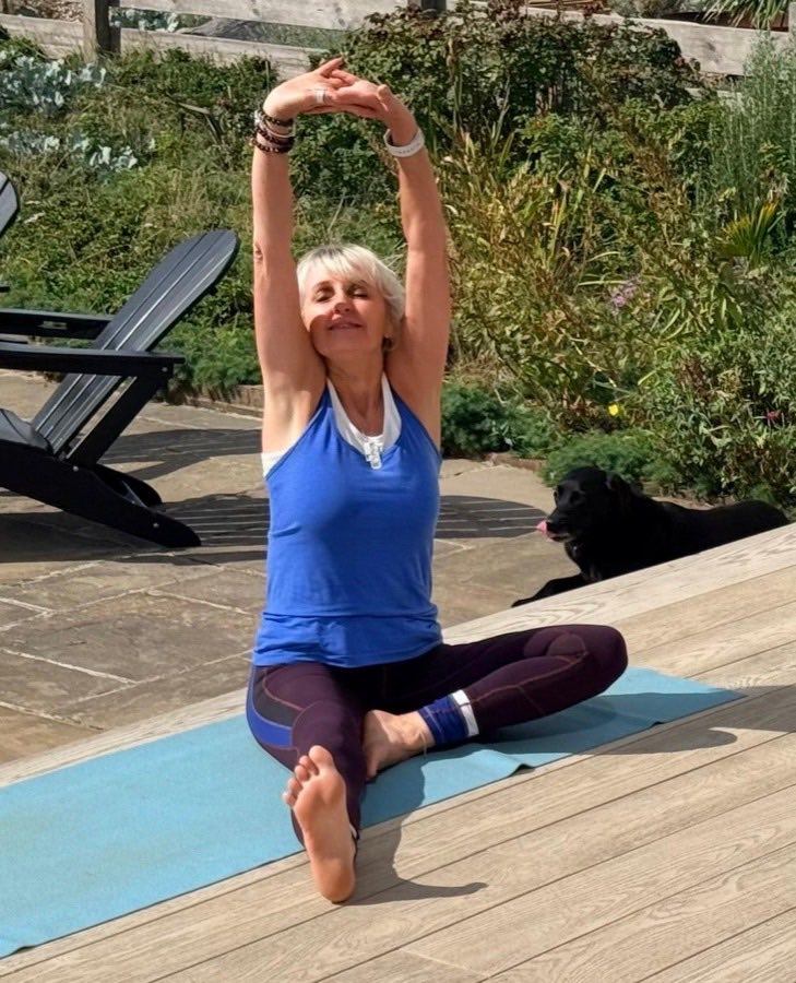 Sally Bayly sitting cross-legged on a mat in the garden, arms stretched overhead, a black labrador behind her.