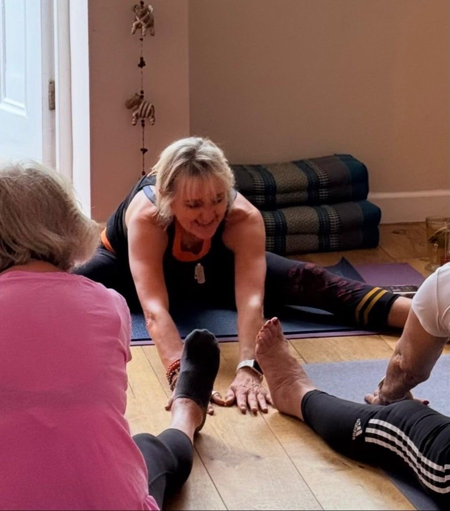 Sally leading a class, reaching forward into a seated stretch with students following.