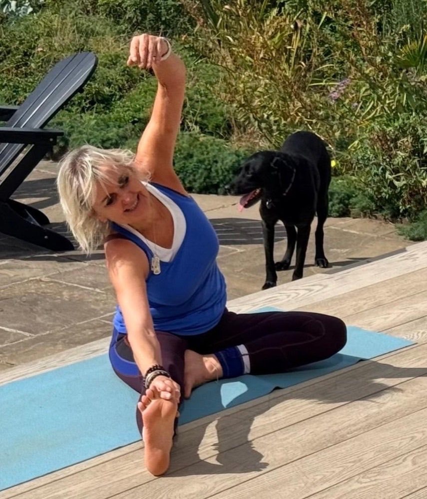 Sally in a seated side stretch on the garden decking, her dog beside her.